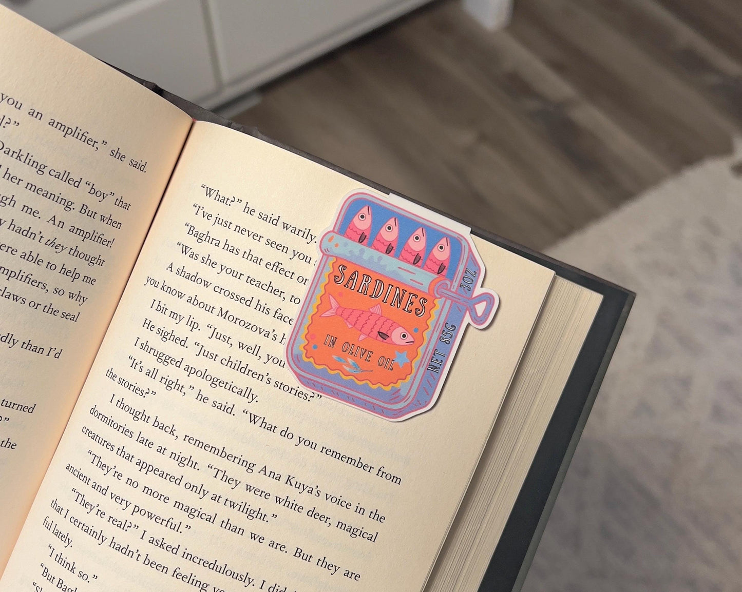 A magnetic bookmark of a pink and orange cartoon style sardine can displayed on a book