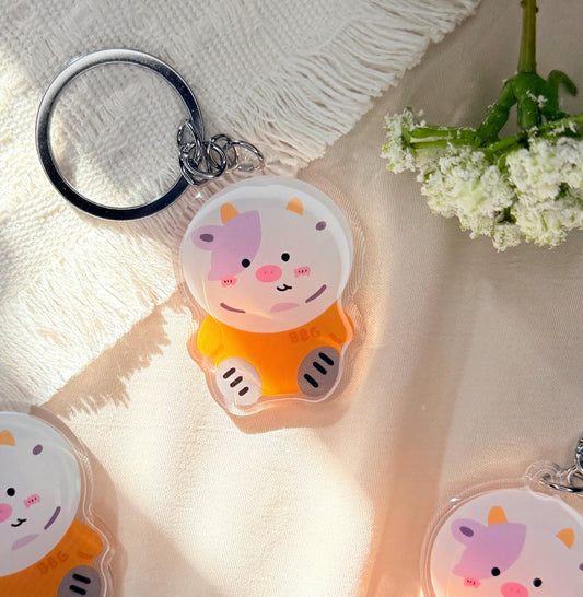 An acrylic keychain of a Kawaii style purple spotted cow dressed in an orange astronaut suit