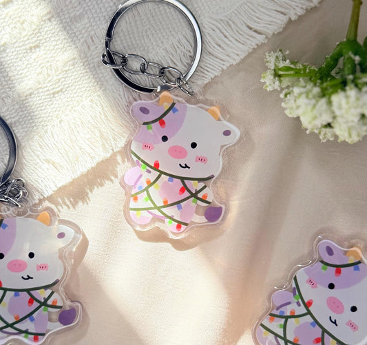 An acrylic keychain of a Kawaii style purple spotted cow wrapped in Christmas lights