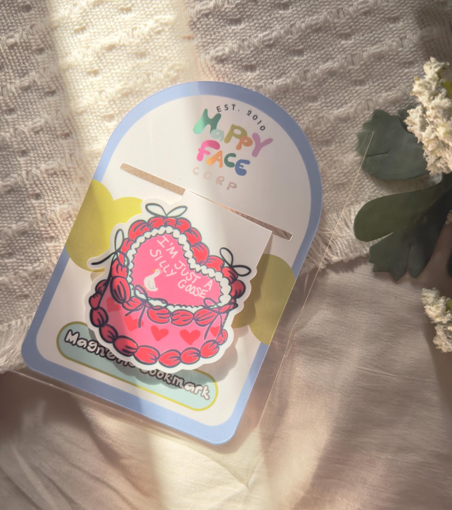 A magnetic bookmark of a pink and red frosted heart shaped cake that has a tiny goose, and it says just a silly goose on the cake