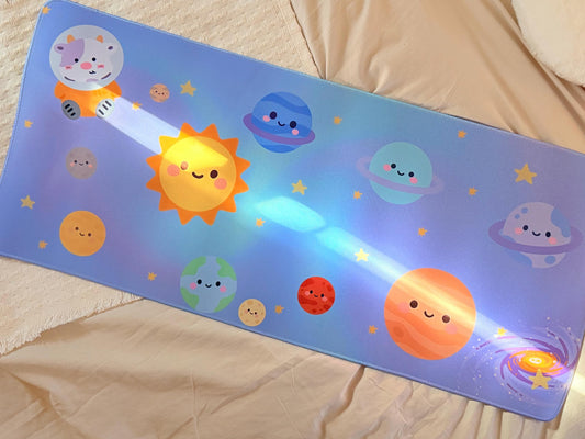 A keyboard sized mouse pad that has a scene of a blue purple space theme with nine planets and the sun in the middle, but they caught purple spotted cow astronaut