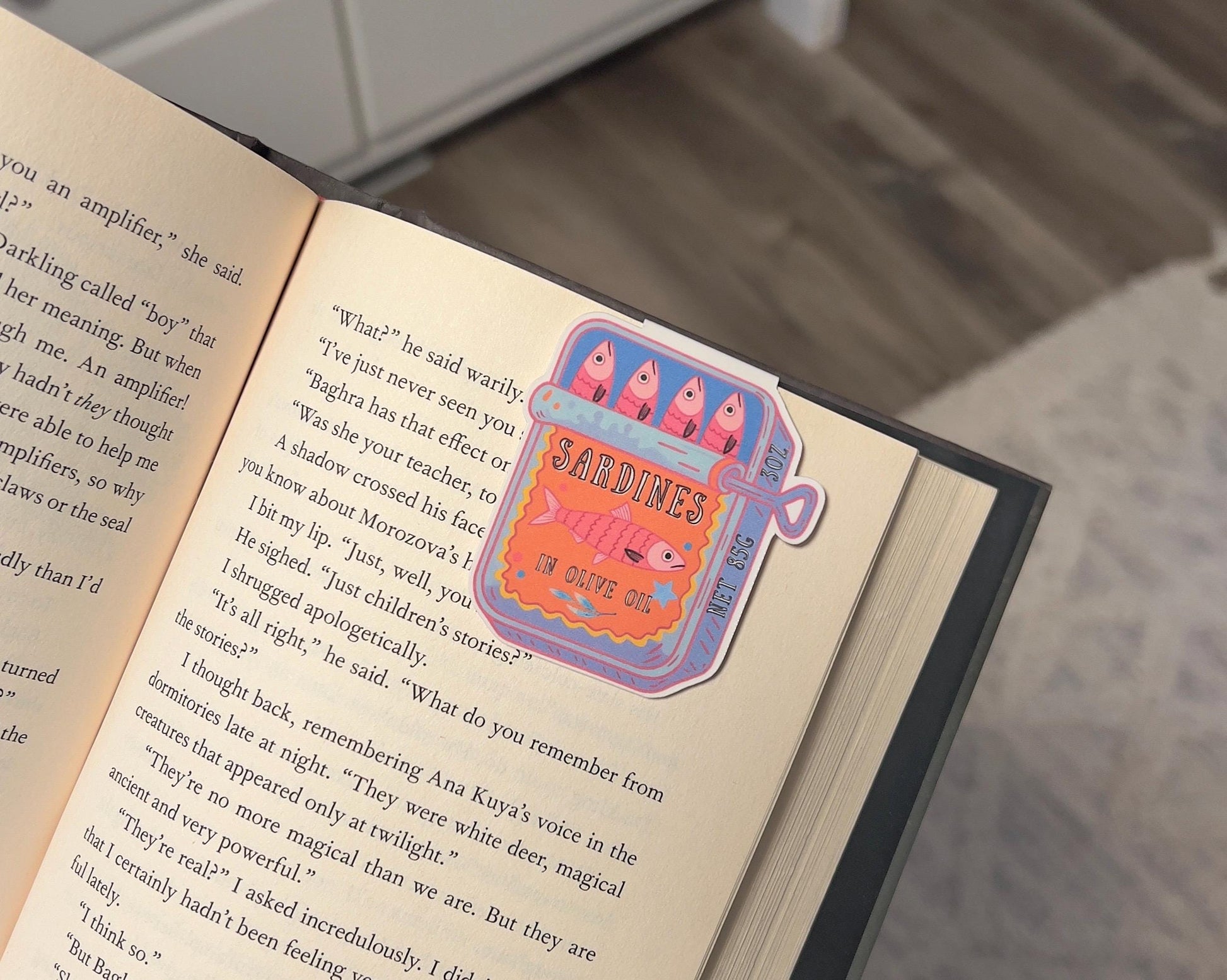 A magnetic bookmark of a pink and orange cartoon style sardine can displayed on a book